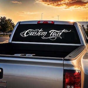 May include: A silver pickup truck features a white vinyl decal applied to the rear window. The decal displays the words Custom Text in a decorative, flowing script font with elegant flourishes extending from the letters across the glass.