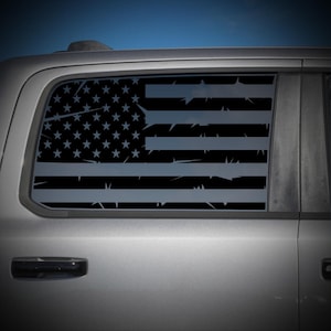May include: A dark gray vinyl decal applied to a vehicle window, featuring a stylized American flag design. The flag incorporates a barbed wire motif integrated into the horizontal stripes and the star field, creating a distressed appearance.