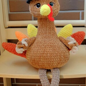 May include: A handmade plush turkey doll, perfect for Thanksgiving. The turkey is brown with colorful orange, yellow, and red tail feathers. It wears a black hat with a green band and has a yellow beak and red wattle. The doll is sitting on a wooden chair.