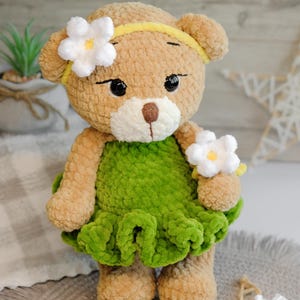 May include: A crocheted teddy bear wearing a green dress with white flowers. The bear has brown fur and black eyes.