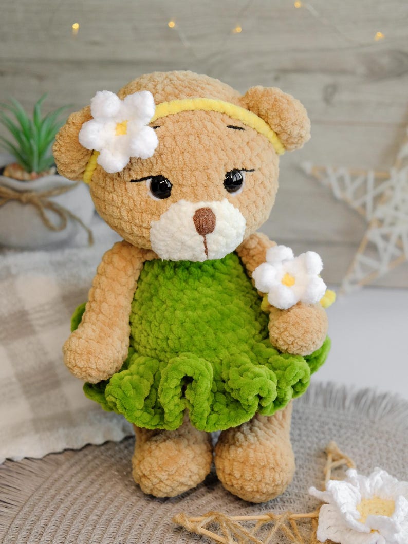 May include: A crocheted teddy bear wearing a green dress with white flowers. The bear has brown fur and black eyes.