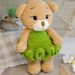 May include: A crocheted plush toy bear wearing a green dress. The bear has brown fur, black eyes, and a white nose. The bear is standing on a light brown surface.