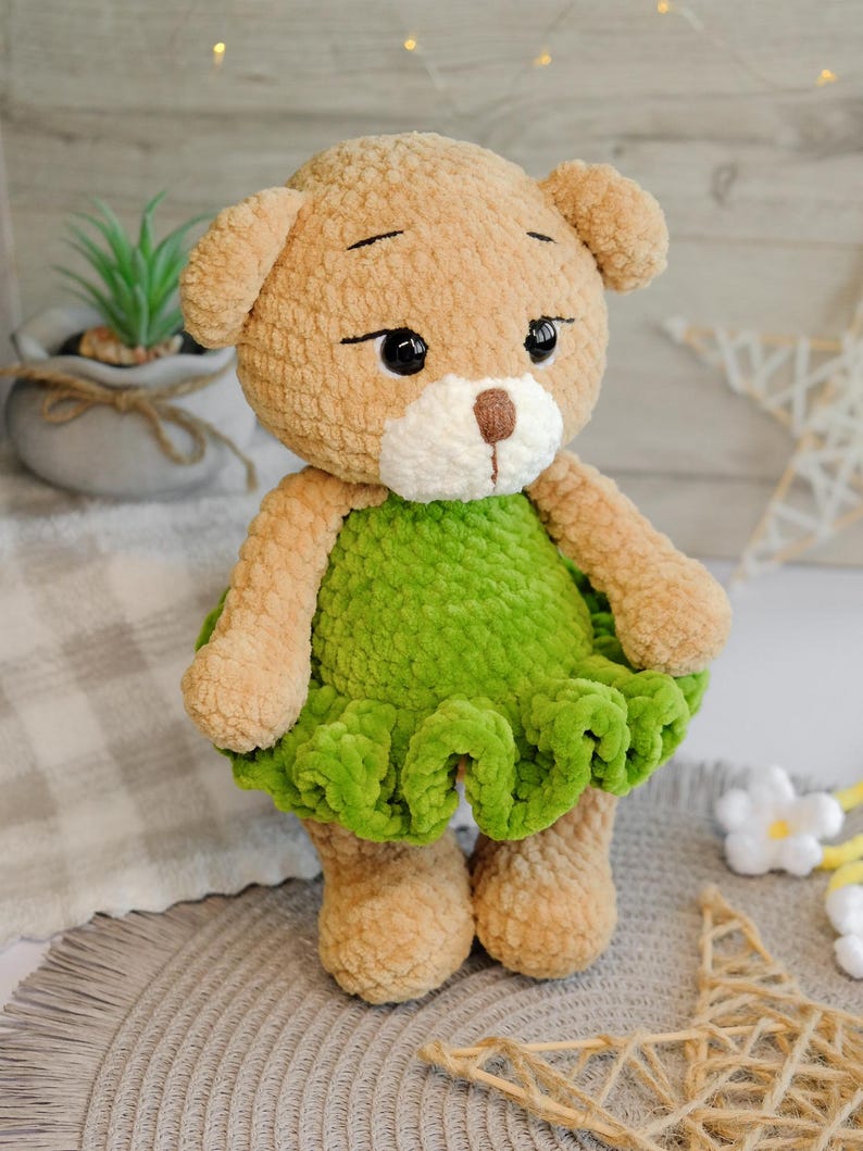 May include: A crocheted plush toy bear wearing a green dress. The bear has brown fur, black eyes, and a white nose. The bear is standing on a light brown surface.