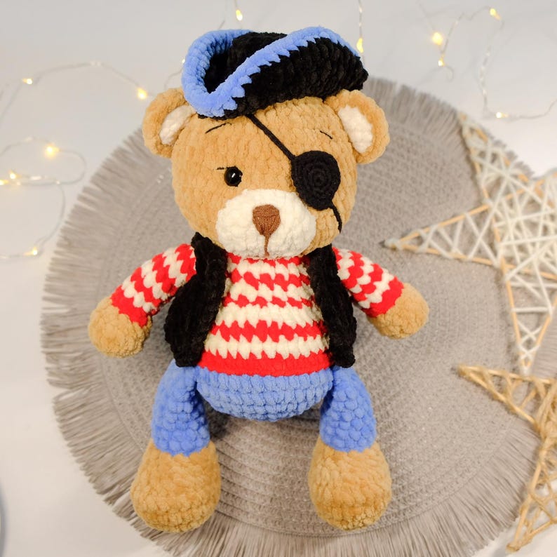 Peut inclure: Un ours en peluche crochet&eacute; brun portant un chapeau de pirate noir et bleu, une chemise ray&eacute;e rouge et blanche, un gilet noir et un cache-&oelig;il bleu.