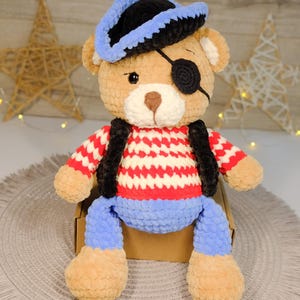Peut inclure: Un ours en peluche crochet&eacute; portant un chapeau de pirate bleu et noir, un cache-&oelig;il et une chemise ray&eacute;e rouge et blanche avec un pantalon bleu.