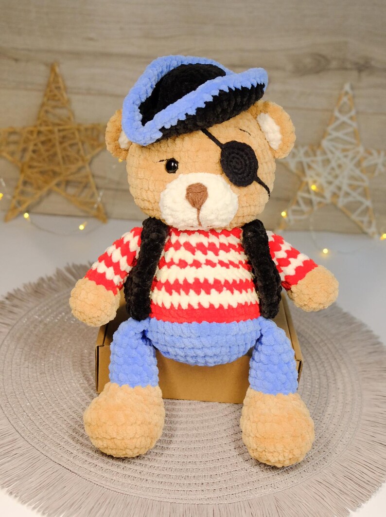 Peut inclure: Un ours en peluche crochet&eacute; portant un chapeau de pirate bleu et noir, un cache-&oelig;il et une chemise ray&eacute;e rouge et blanche avec un pantalon bleu.