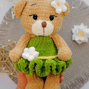 May include: A crocheted brown teddy bear wearing a green dress with a white flower on the dress and a white flower in its hair.