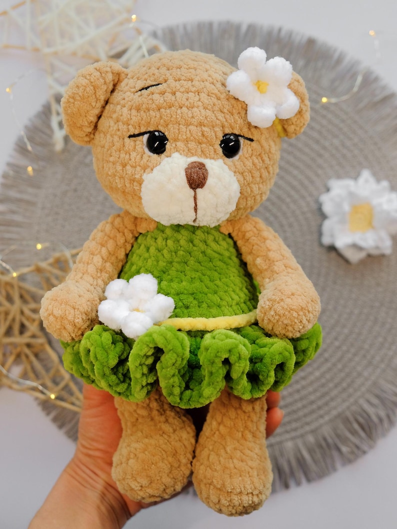 May include: A crocheted brown teddy bear wearing a green dress with a white flower on the dress and a white flower in its hair.