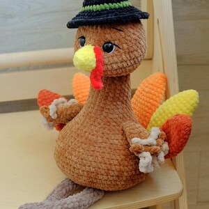 May include: A brown crocheted turkey doll with a black hat featuring a green band. The turkey has a yellow beak, red wattle, and colorful tail feathers in orange, yellow, and red. It is sitting on a wooden surface.