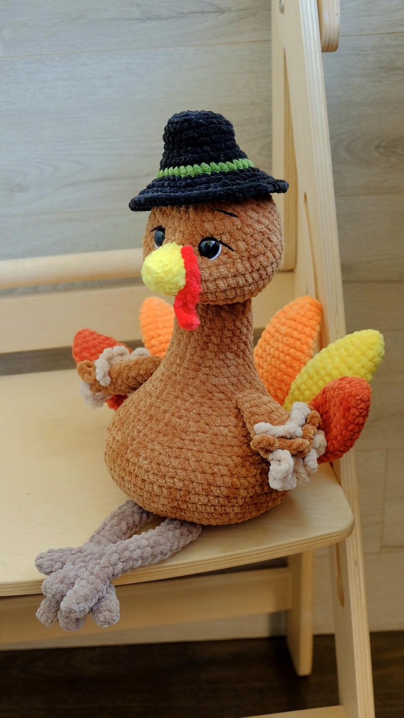 May include: A brown crocheted turkey doll with a black hat featuring a green band. The turkey has a yellow beak, red wattle, and colorful tail feathers in orange, yellow, and red. It is sitting on a wooden surface.