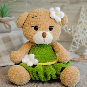 May include: A crocheted teddy bear wearing a green dress with white flowers. The bear has brown fur and black eyes.