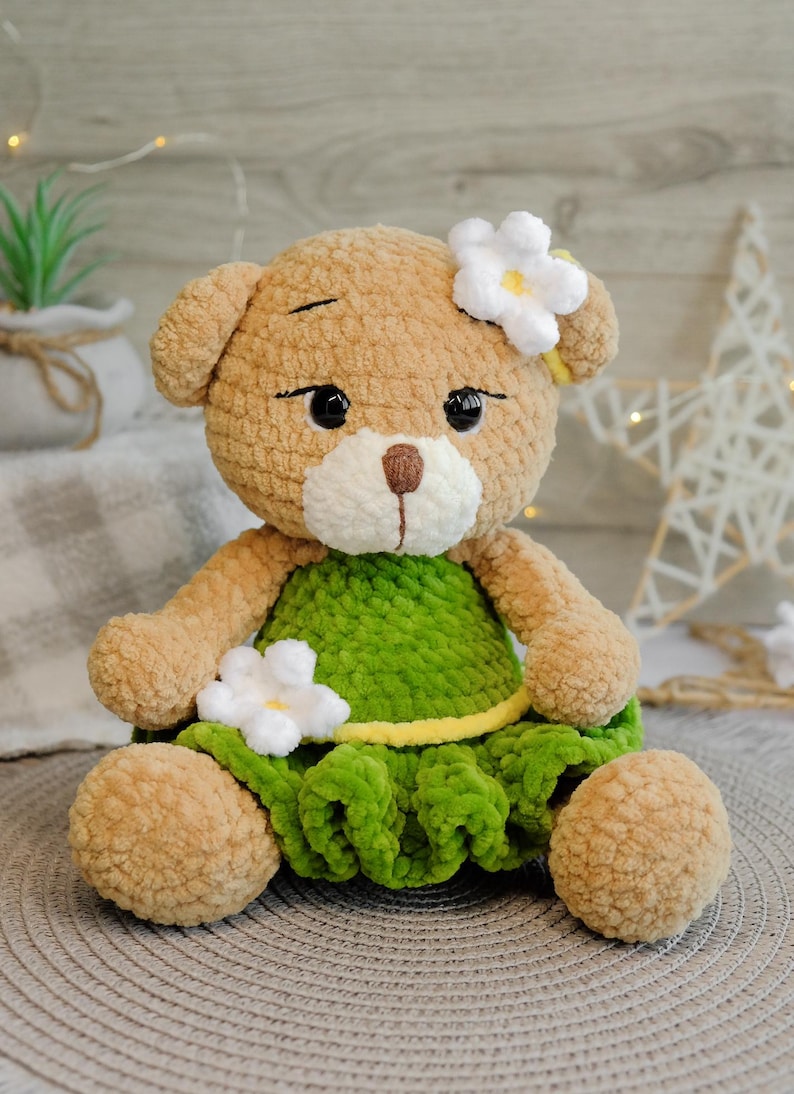 May include: A crocheted teddy bear wearing a green dress with white flowers. The bear has brown fur and black eyes.