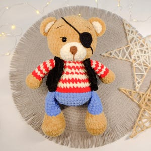 Peut inclure: Un ours en peluche crochet&eacute; portant une chemise ray&eacute;e rouge et blanche, un gilet noir et un jean bleu. L'ours a un cache-&oelig;il brun et noir.