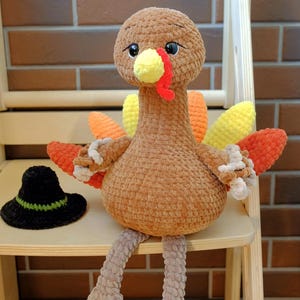 May include: A handmade, plush turkey with a brown body, colorful tail feathers, and a yellow beak and wattle. It sits on a wooden chair next to a small black hat with a green brim. The turkey is a cute, decorative item.