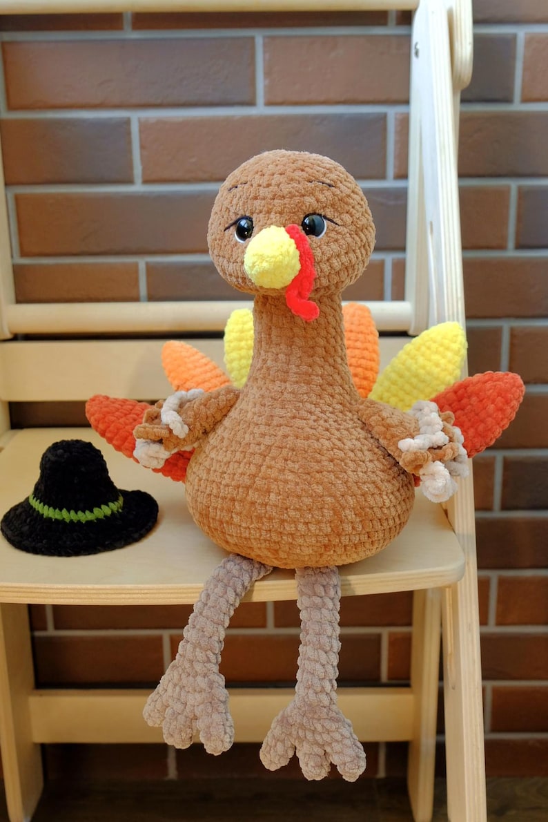 May include: A handmade, plush turkey with a brown body, colorful tail feathers, and a yellow beak and wattle. It sits on a wooden chair next to a small black hat with a green brim. The turkey is a cute, decorative item.