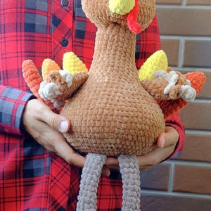 May include: A handmade, plush turkey toy with a brown body, head, and neck. It has a yellow beak, red wattle, and colorful tail feathers in shades of orange, yellow, and white. The turkey has gray legs and is held in front of a red plaid shirt.