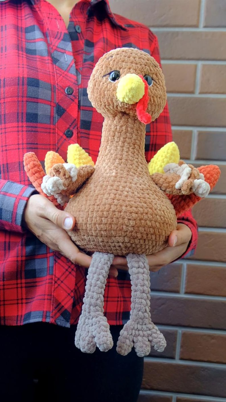 May include: A handmade, plush turkey toy with a brown body, head, and neck. It has a yellow beak, red wattle, and colorful tail feathers in shades of orange, yellow, and white. The turkey has gray legs and is held in front of a red plaid shirt.
