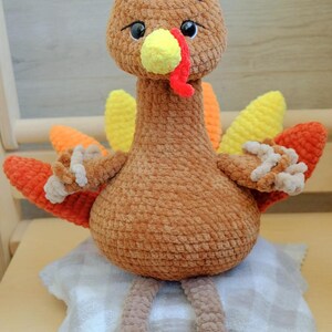 May include: A handmade, plush turkey toy with a brown body, head, and legs. It has a yellow beak and wattle, and colorful orange and yellow tail feathers. The turkey is sitting on a small, checkered pillow.