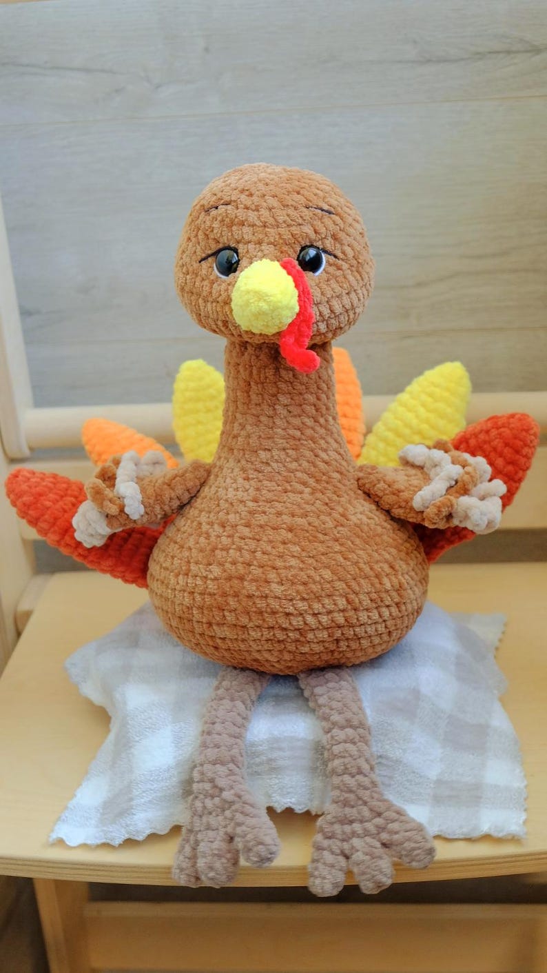 May include: A handmade, plush turkey toy with a brown body, head, and legs. It has a yellow beak and wattle, and colorful orange and yellow tail feathers. The turkey is sitting on a small, checkered pillow.