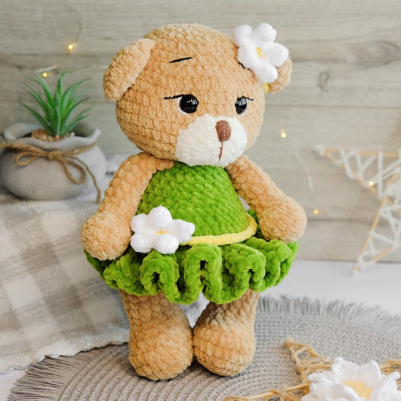 May include: A crocheted teddy bear wearing a green dress with a yellow trim and white flowers. The bear has brown fur and black eyes.