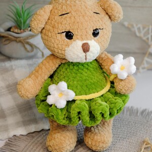 May include: A crocheted teddy bear wearing a green dress with white flowers. The bear has brown fur and black eyes.