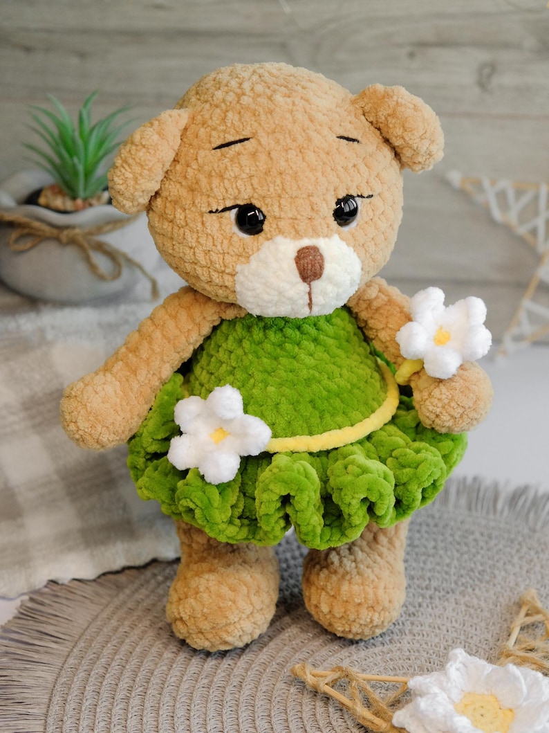 May include: A crocheted teddy bear wearing a green dress with white flowers. The bear has brown fur and black eyes.