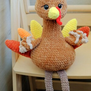 May include: A handmade plush turkey toy with a brown body, head, and legs. The turkey has yellow, orange, and red wings, a yellow beak, and a red wattle. The turkey is sitting on a wooden chair.