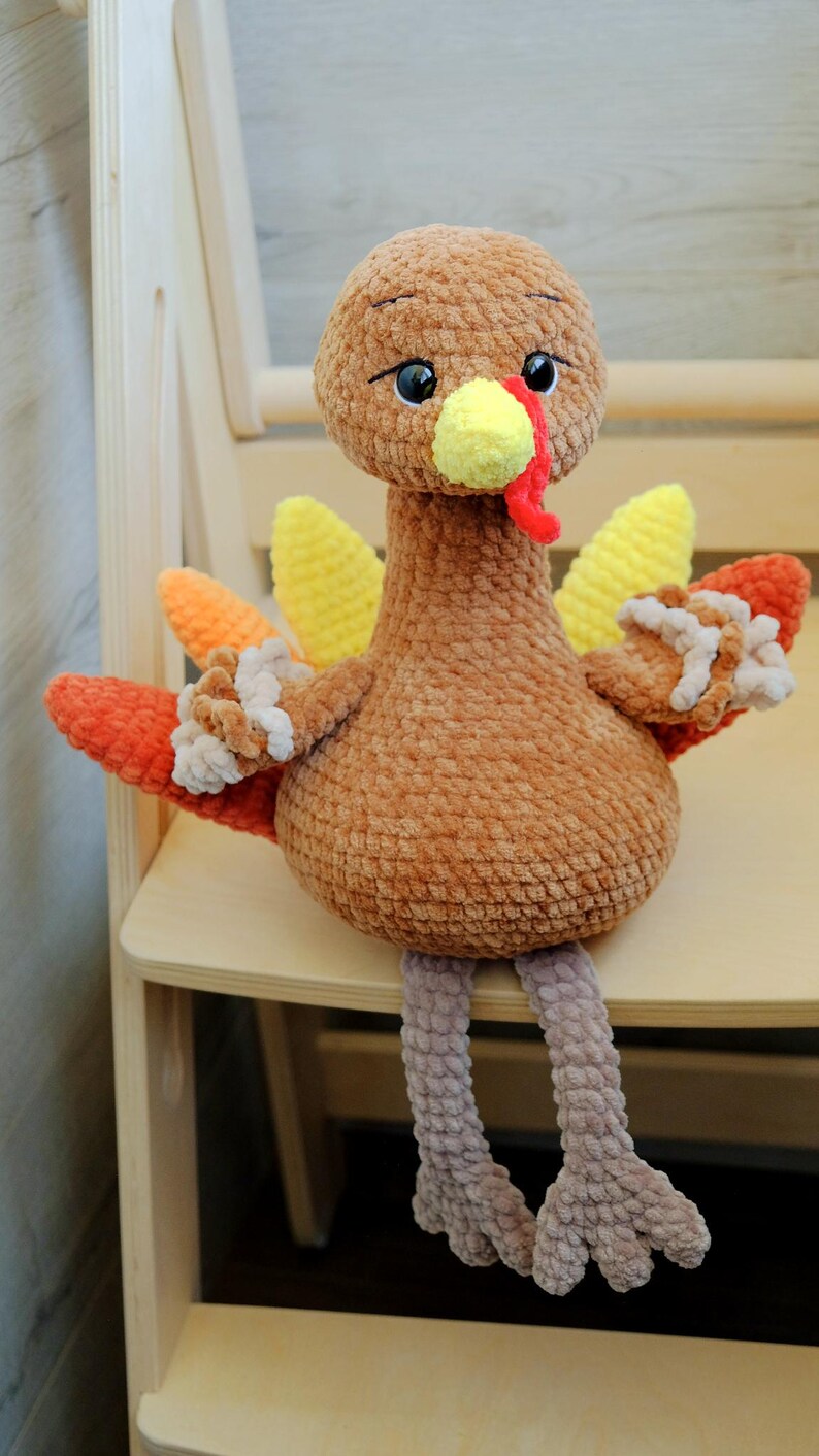 May include: A handmade plush turkey toy with a brown body, head, and legs. The turkey has yellow, orange, and red wings, a yellow beak, and a red wattle. The turkey is sitting on a wooden chair.