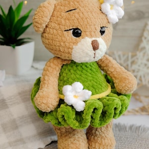 May include: A crocheted brown teddy bear wearing a green dress with white flowers and a yellow belt. The bear has black eyes and a white nose.