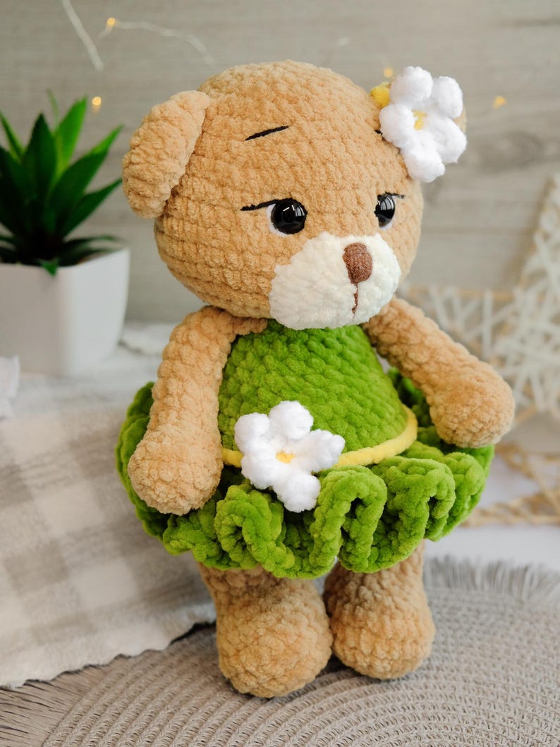May include: A crocheted brown teddy bear wearing a green dress with white flowers and a yellow belt. The bear has black eyes and a white nose.