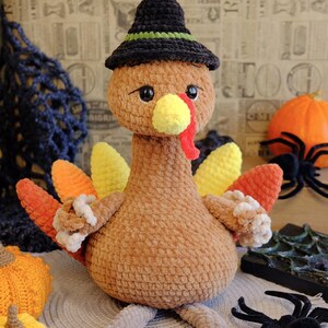 May include: A handmade, brown crochet turkey wearing a black witch hat with a green band. The turkey has yellow, orange, and red tail feathers and a yellow beak. Decorative spiders and pumpkins are in the background.