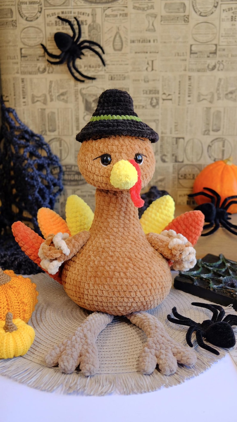 May include: A handmade, brown crochet turkey wearing a black witch hat with a green band. The turkey has yellow, orange, and red tail feathers and a yellow beak. Decorative spiders and pumpkins are in the background.