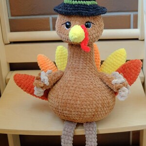 May include: A handmade crochet turkey doll with a black hat with a green band. The turkey has a brown body, yellow beak, red wattle, and colorful tail feathers in shades of orange and yellow. It is sitting on a wooden chair.