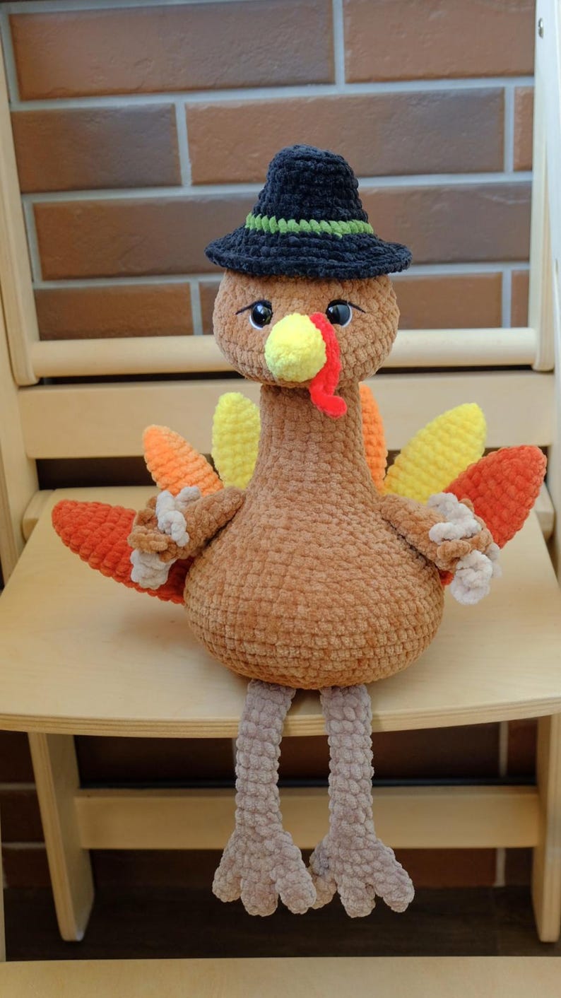 May include: A handmade crochet turkey doll with a black hat with a green band. The turkey has a brown body, yellow beak, red wattle, and colorful tail feathers in shades of orange and yellow. It is sitting on a wooden chair.