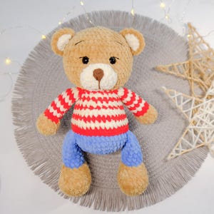 Peut inclure: Un ours en peluche crochet&eacute; portant un pull ray&eacute; rouge et blanc et un jean bleu. L'ours est assis sur un tapis gris &agrave; franges.