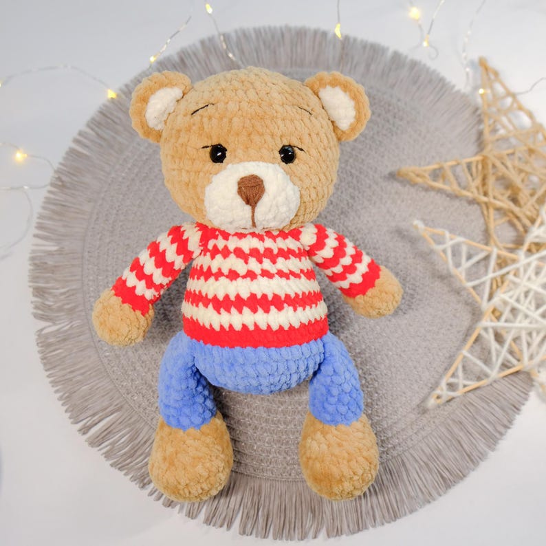 Peut inclure: Un ours en peluche crochet&eacute; portant un pull ray&eacute; rouge et blanc et un jean bleu. L'ours est assis sur un tapis gris &agrave; franges.