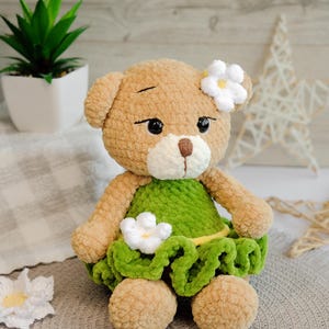 May include: A crocheted teddy bear wearing a green dress with white flowers. The bear has brown fur and black eyes.