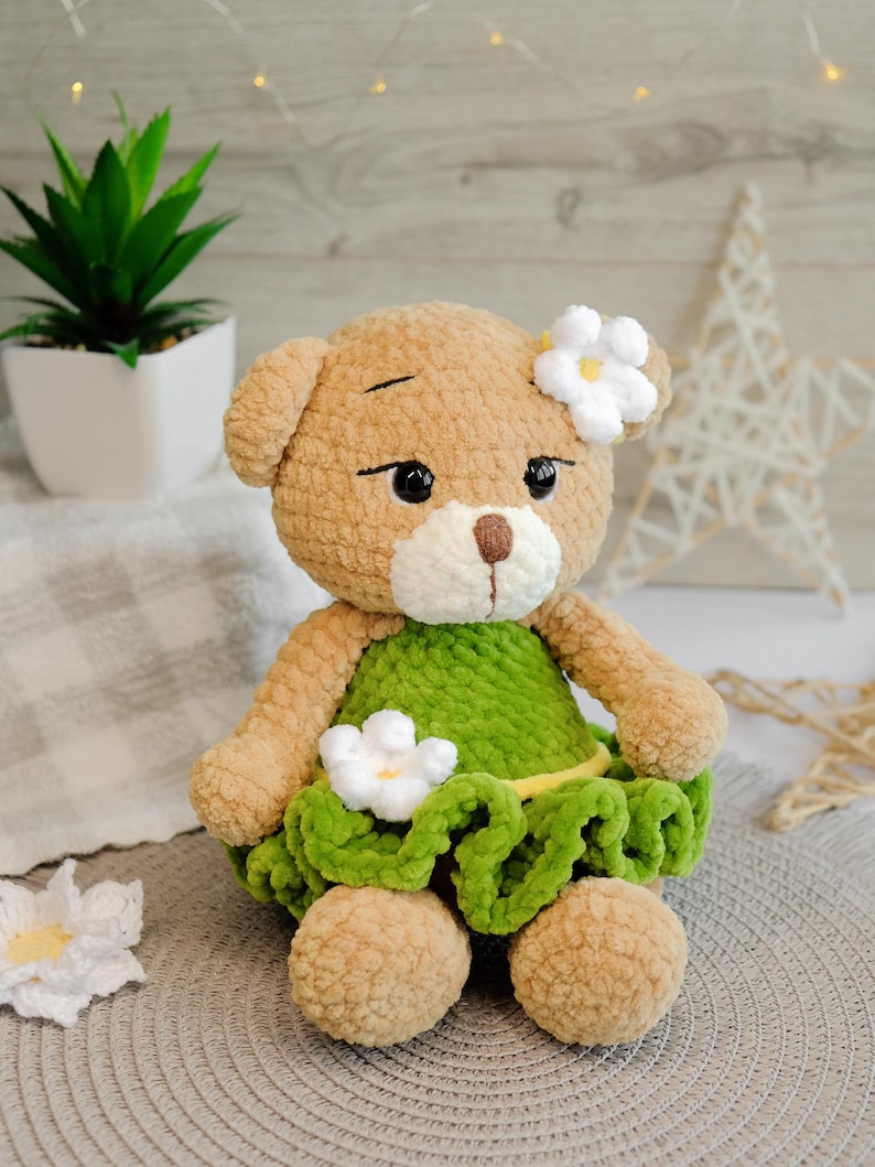 May include: A crocheted teddy bear wearing a green dress with white flowers. The bear has brown fur and black eyes.