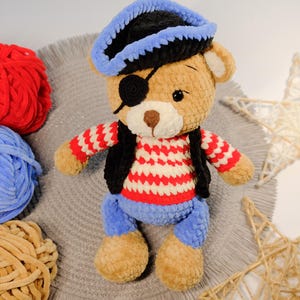 Peut inclure: Un ours en peluche crochet&eacute; portant un chapeau de pirate bleu et noir, une chemise ray&eacute;e rouge et blanche, un gilet noir et un cache-&oelig;il bleu. L'ours est assis sur une surface grise avec des pelotes de laine en arri&egrave;re-plan.
