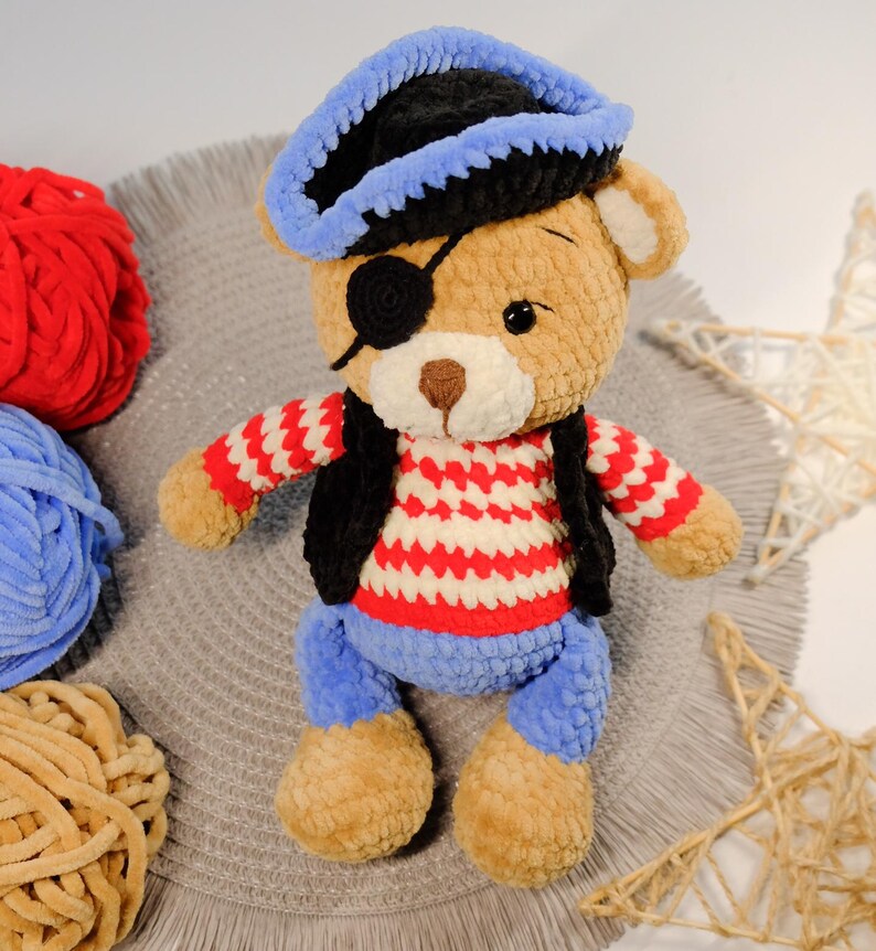 Peut inclure: Un ours en peluche crochet&eacute; portant un chapeau de pirate bleu et noir, une chemise ray&eacute;e rouge et blanche, un gilet noir et un cache-&oelig;il bleu. L'ours est assis sur une surface grise avec des pelotes de laine en arri&egrave;re-plan.