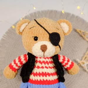 Peut inclure: Un ours en peluche crochet&eacute; marron portant un pull ray&eacute; rouge et blanc, un gilet noir et un cache-&oelig;il noir. L'ours est assis sur une surface duveteuse grise et blanche.