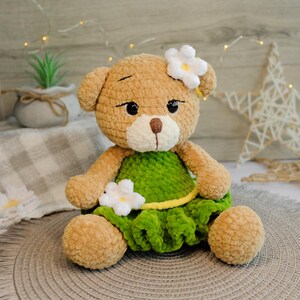 May include: A crocheted teddy bear wearing a green dress with white flowers and a yellow sash. The bear has brown fur and black eyes.