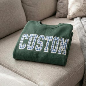 May include: A folded, forest green sweatshirt with the word "CUSTOM" in large, white, block letters. The letters are filled with a blue and white gingham pattern. The sweatshirt is resting on a light gray couch.