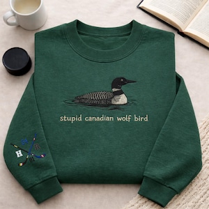 Puede incluir: Sudadera verde oscuro con un bordado de un somorgujo nadando en agua. El texto "stupid canadian wolf bird" está debajo del ave. La manga izquierda tiene palos de hockey bordados y las letras "H" y "R".