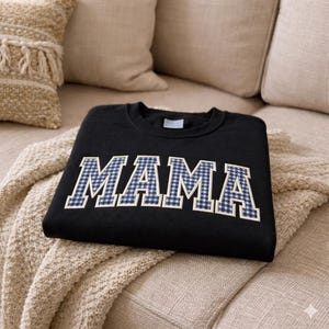 May include: Black sweatshirt with the word "MAMA" embroidered in white letters with a blue and white gingham pattern. The sweatshirt is folded on a beige knitted blanket, with beige pillows in the background.