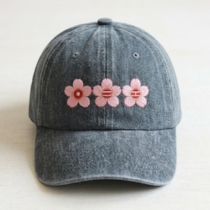 May include: A gray baseball cap with three embroidered pink flowers across the front. The flowers have red and white details. The cap has a curved brim and a button on top.