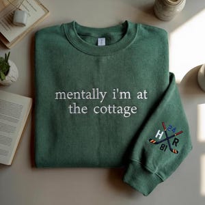 May include: A green sweatshirt with the embroidered text "mentally I'm at the cottage" in white. The sleeve features crossed hockey sticks and the letters "H", "R", and numbers "24" and "81".