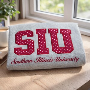 May include: A gray rectangular pillow with the letters "SIU" in red with white polka dots. The words "Southern Illinois University" are embroidered in red below the letters. The pillow is on a wooden surface.