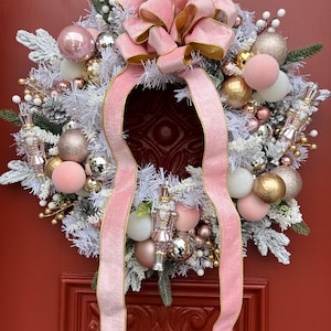 May include: A festive Christmas wreath with a pink and gold color scheme. The wreath features a large pink bow, pink and gold ornaments, white frosted branches, and decorative nutcrackers. The wreath is displayed on a red door.