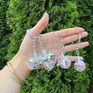 May include: Silver keychains with disco ball and star charms. The charms have the letters "MJ" and "T" on them. The disco ball charms are pink and silver. The background is green.